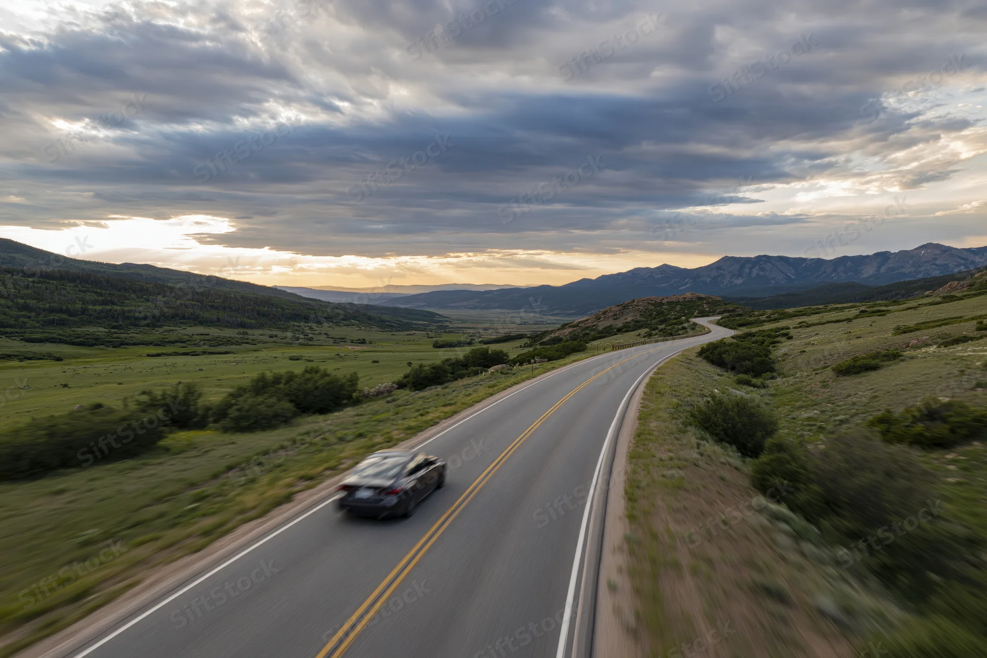 Dark sedan driving away on winding two-lane highway through rural landscape with mountains and cloudy sky at dusk