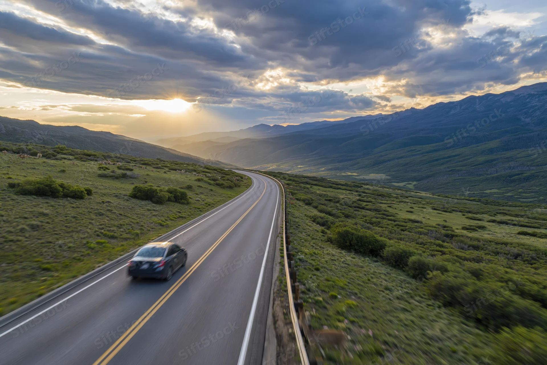 Dark sedan driving away on winding two-lane highway through mountain valley at sunset