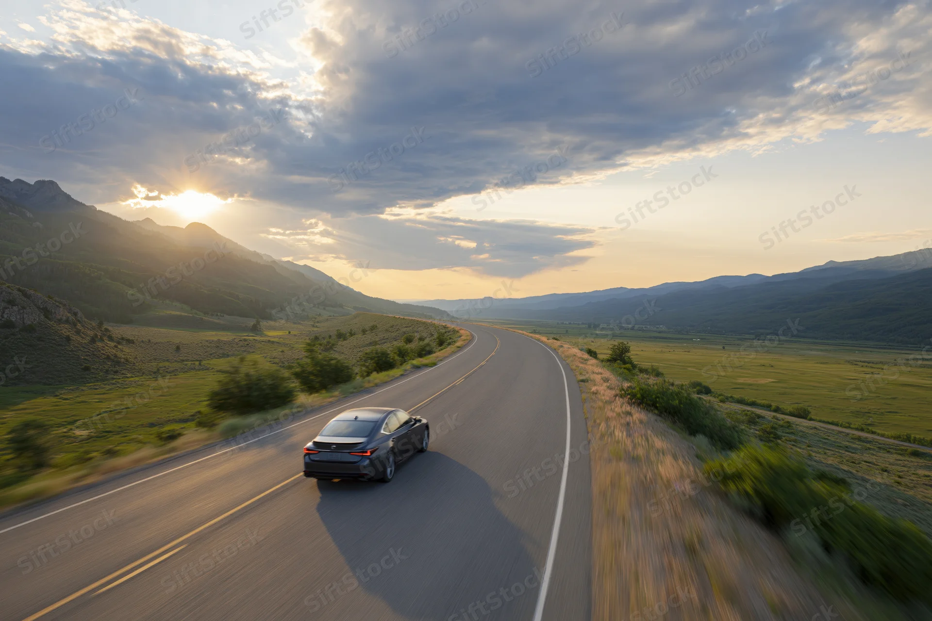 Dark sedan driving away on a winding two-lane highway through a mountain valley at sunset
