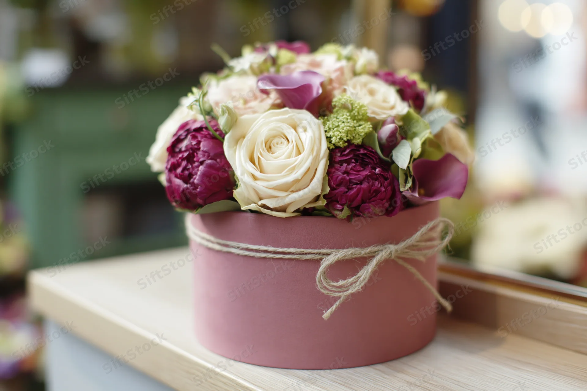 Floral arrangement of cream roses and deep magenta peonies in a pink box tied with twine on a wooden shelf