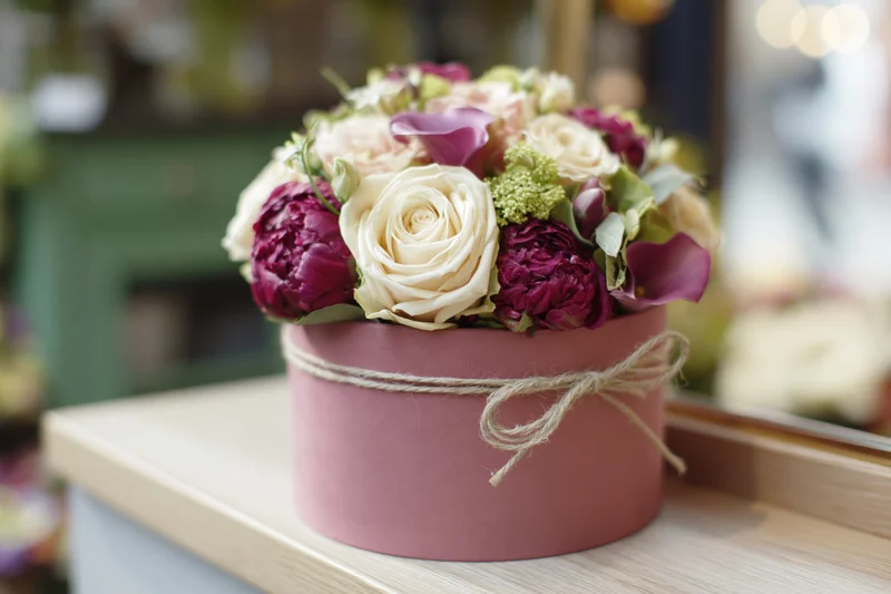 Floral arrangement of cream roses and deep magenta peonies in a pink box tied with twine on a wooden shelf