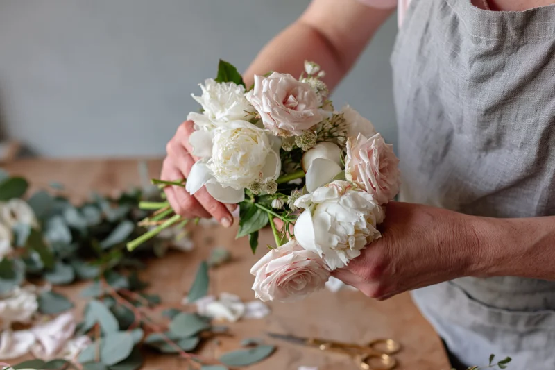 Florist arranging a small bridal bouquet of white peonies and roses on a table with eucalyptus and scissors