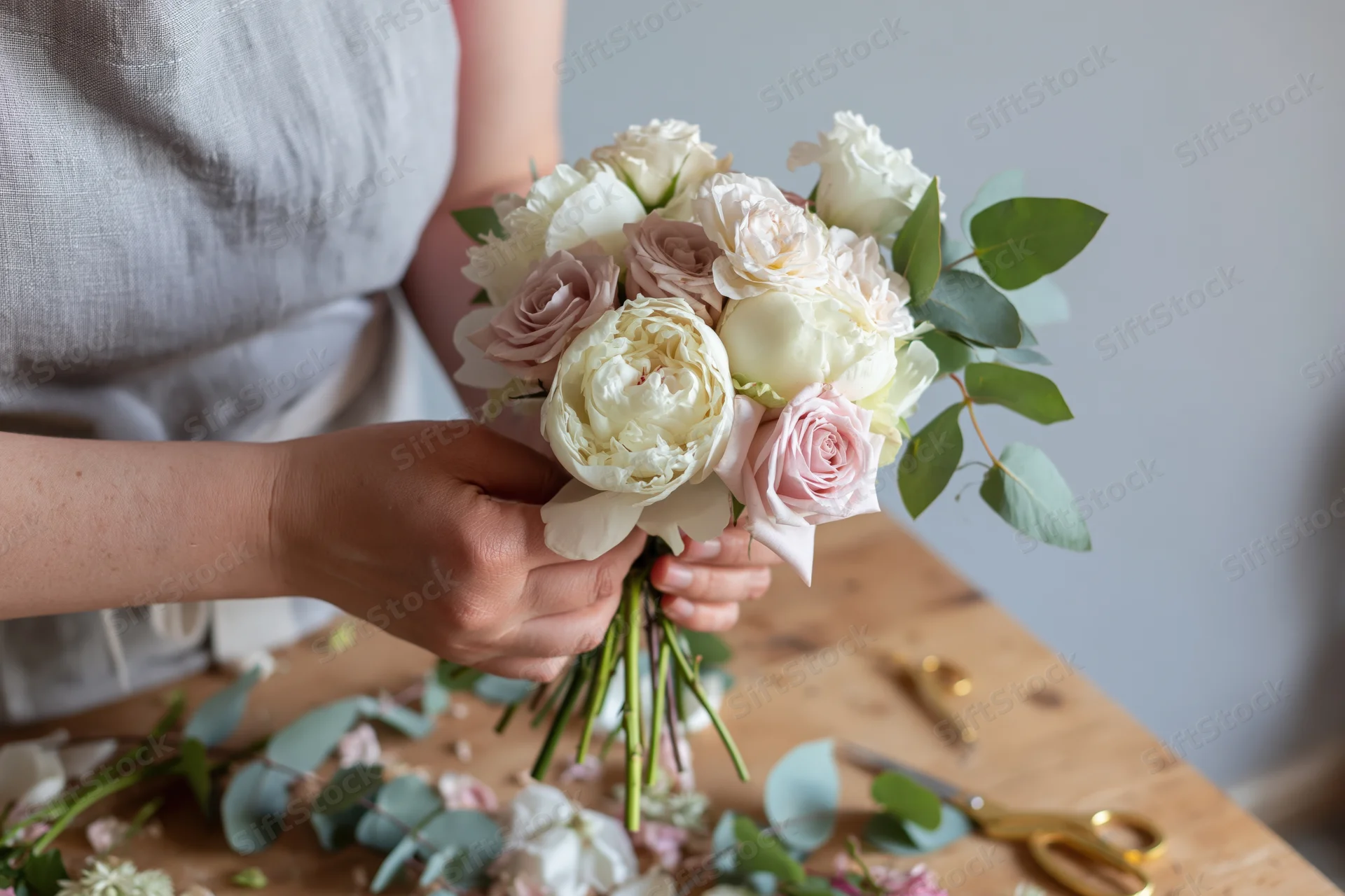 Woman holding a small bridal bouquet of white peonies and roses with eucalyptus on a wooden table