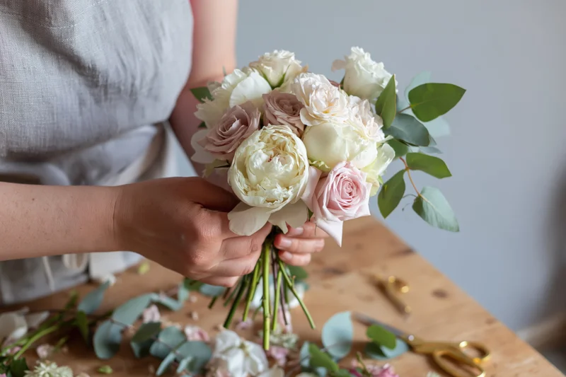 Woman holding a small bridal bouquet of white peonies and roses with eucalyptus on a wooden table