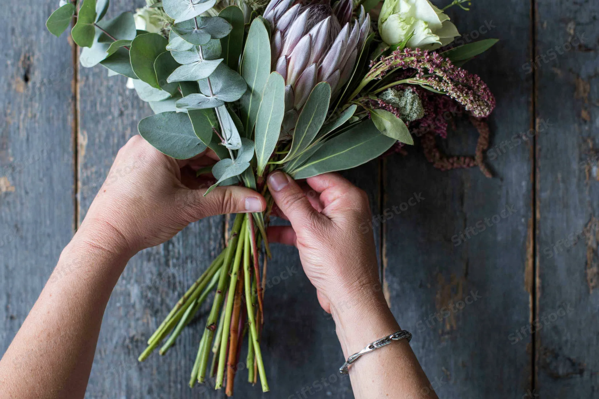 Hands arranging a mixed floral bouquet with protea on a wooden surface