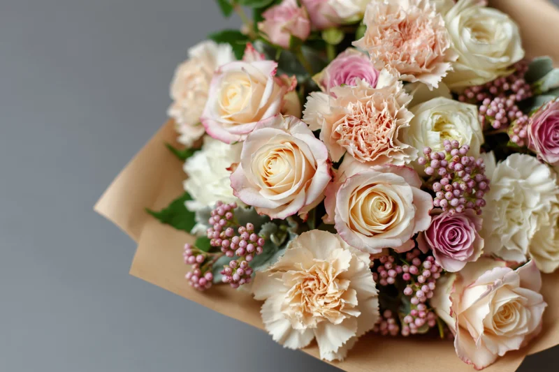 Close-up of a bouquet of cream and blush pink roses and carnations wrapped in kraft paper on a gray surface