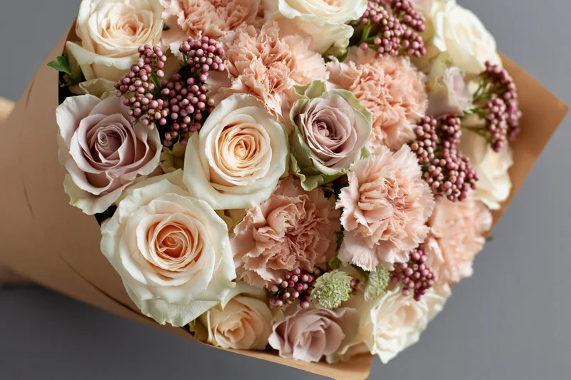 Close-up of a bouquet of cream and blush pink roses and carnations wrapped in brown paper against a gray background