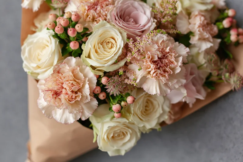 Closeup of a bouquet of cream and blush pink roses and carnations wrapped in kraft paper on a gray surface