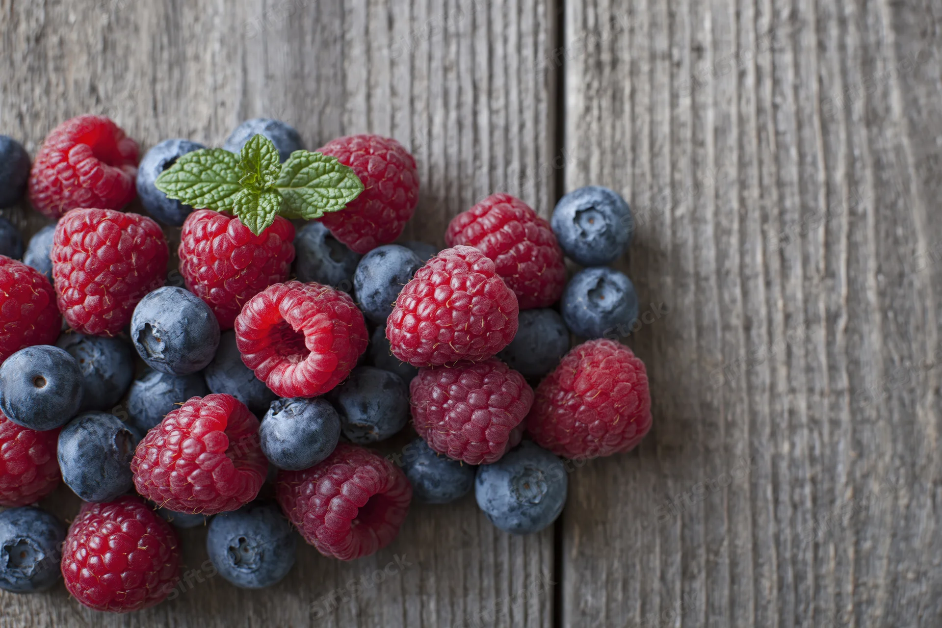 Raspberries and blueberries scattered on a weathered wooden surface with a mint leaf