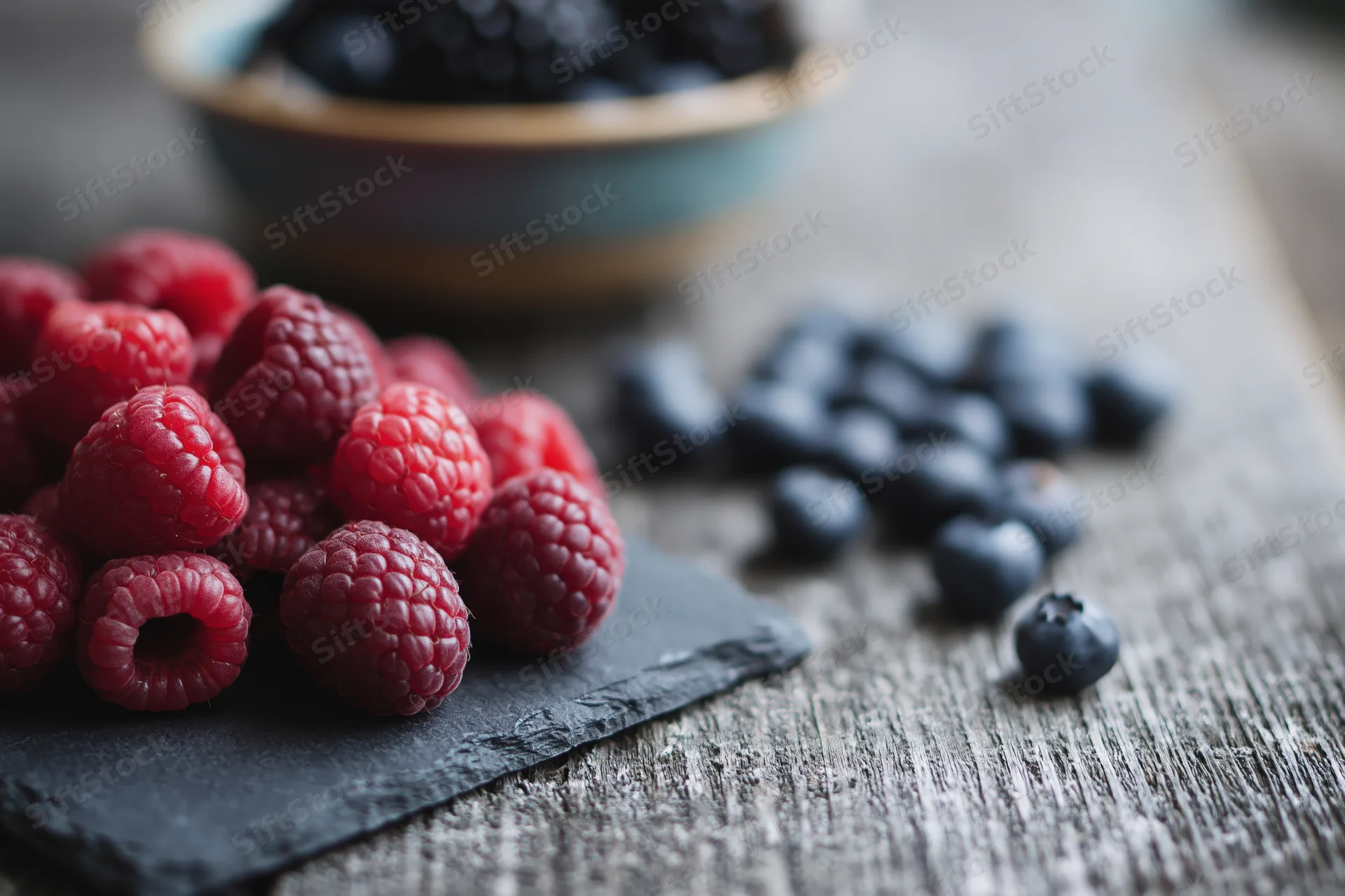 Fresh raspberries piled on a dark slate board beside loose blueberries on a wooden table