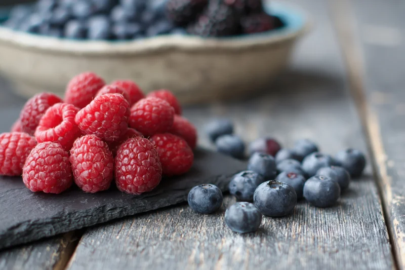 Fresh raspberries piled on a dark slate board beside loose blueberries on a wooden table