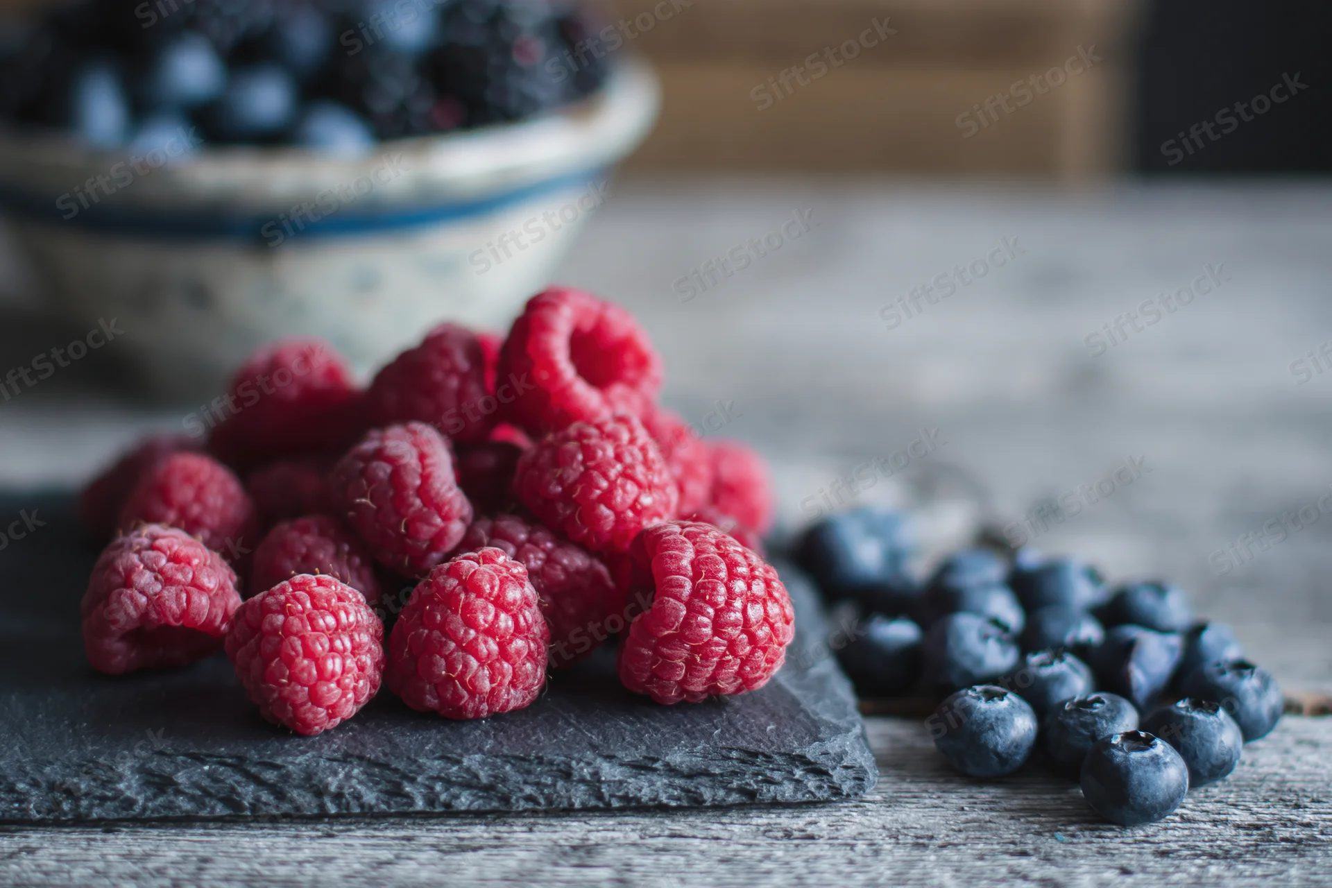 Fresh raspberries piled on a dark slate board beside loose blueberries on a wooden table