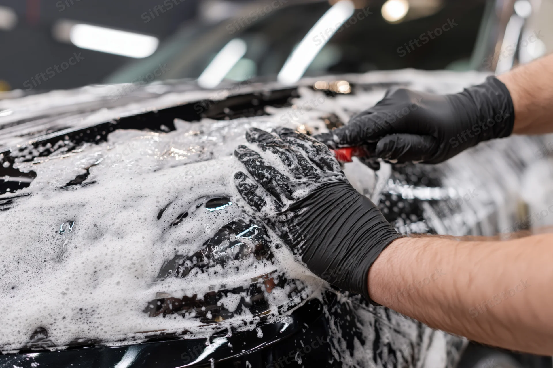 Hands in black nitrile gloves scrubbing a soapy car headlight in a garage