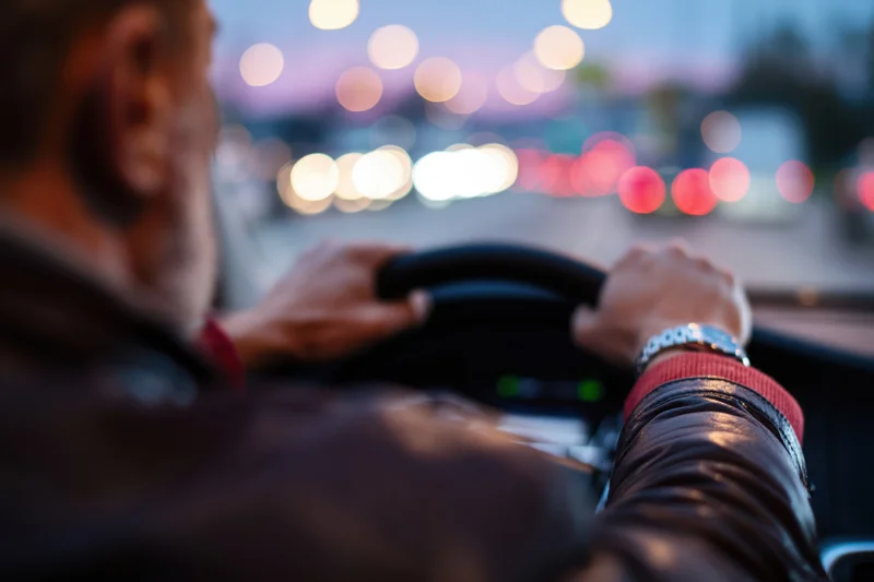 Man driving a car at twilight with hands on steering wheel, city lights blurred in background