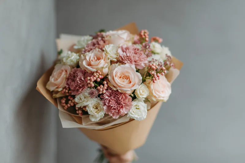 Hand holding a bouquet of pale pink roses and dusty pink carnations wrapped in kraft paper against a gray wall