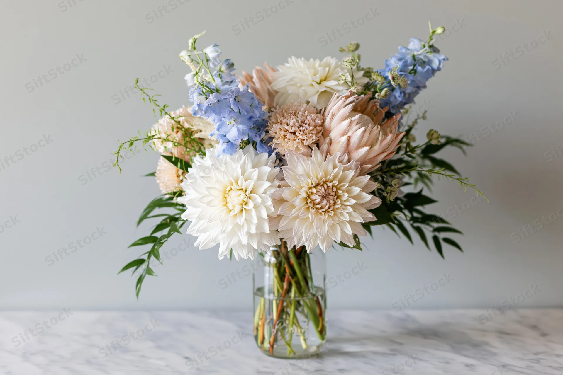 Mixed floral bouquet with dahlias and delphiniums in a clear glass jar on a marble table