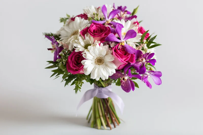 Mixed flower bouquet with roses, orchids and daisies tied with ribbon against white background