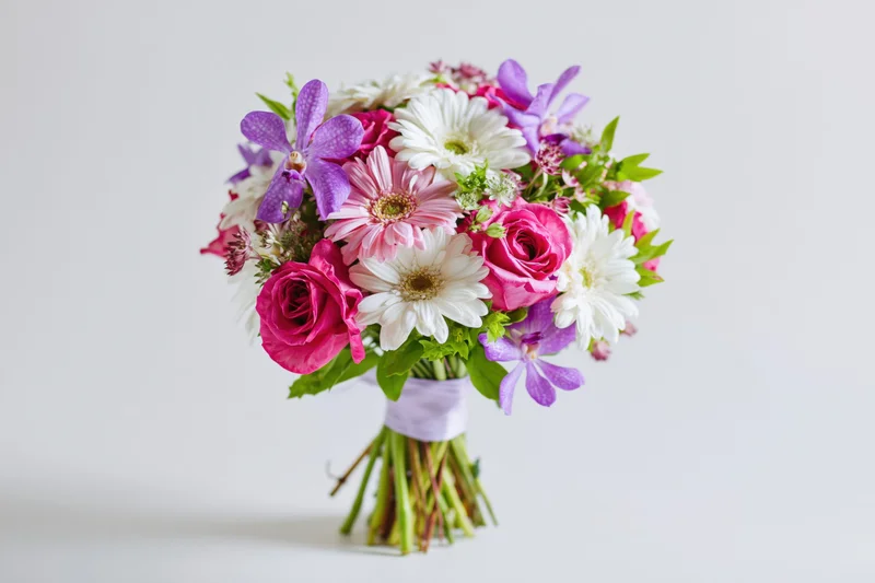 Mixed flower bouquet with pink roses, purple orchids and white daisies wrapped in ribbon against white background