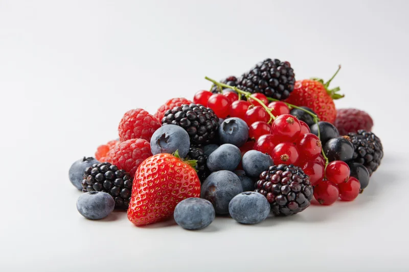 A pile of mixed fresh berries including strawberries, blueberries, blackberries, raspberries and redcurrants on a white surface