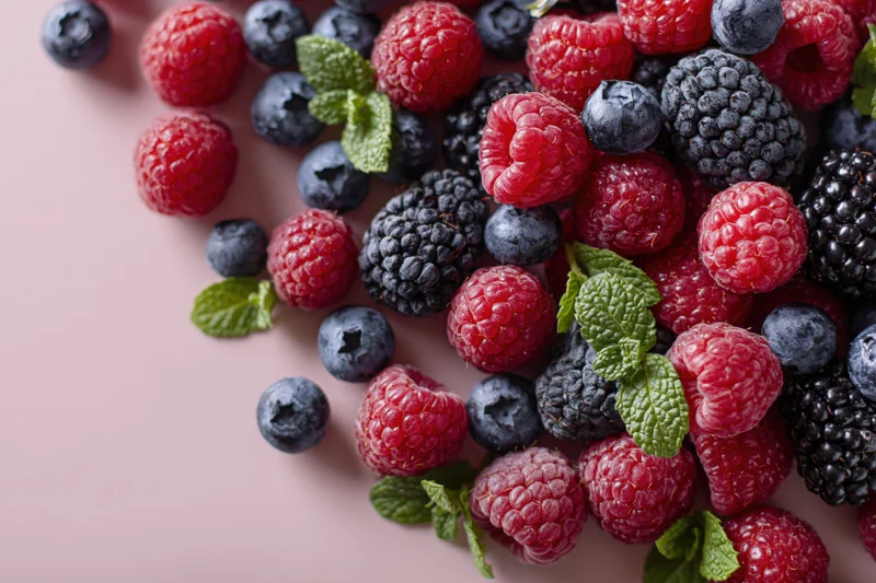 Fresh raspberries blueberries and blackberries with mint leaves arranged on pink surface