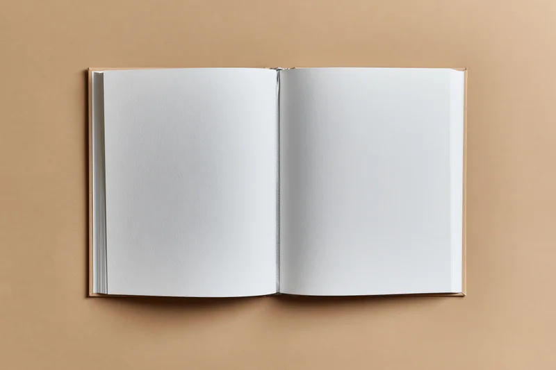 Open hardcover book with blank white pages lying flat on beige surface