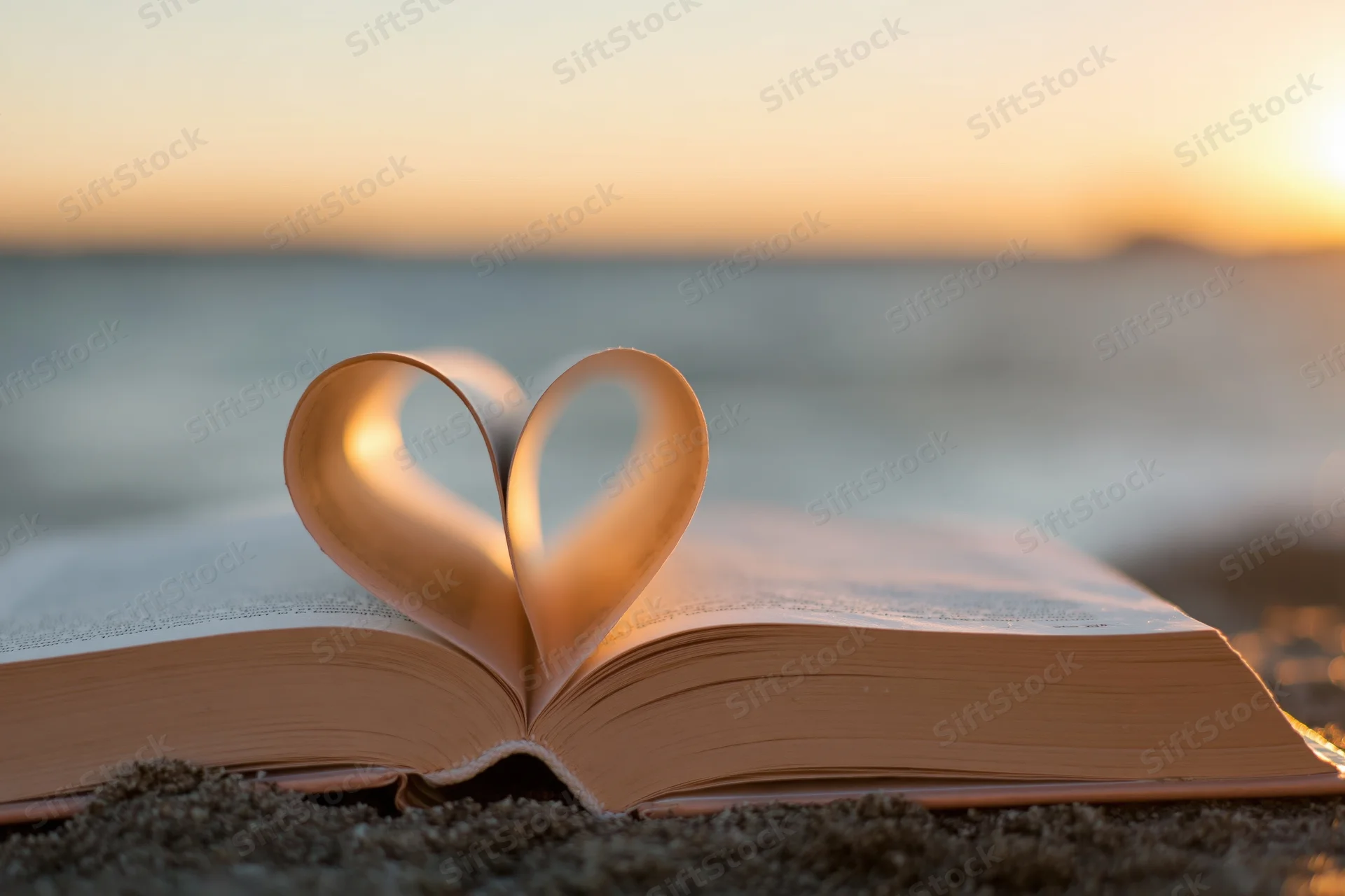 Open hardcover book with pages folded into heart shape on sandy beach at sunset