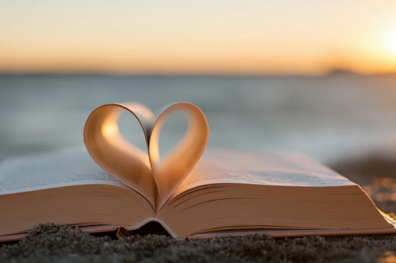 Open hardcover book with pages folded into heart shape on sandy beach at sunset