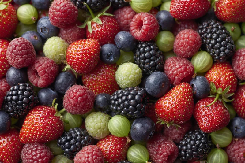 Overhead close-up of a dense pile of mixed fresh berries including strawberries, raspberries, blueberries, blackberries, and gooseberries