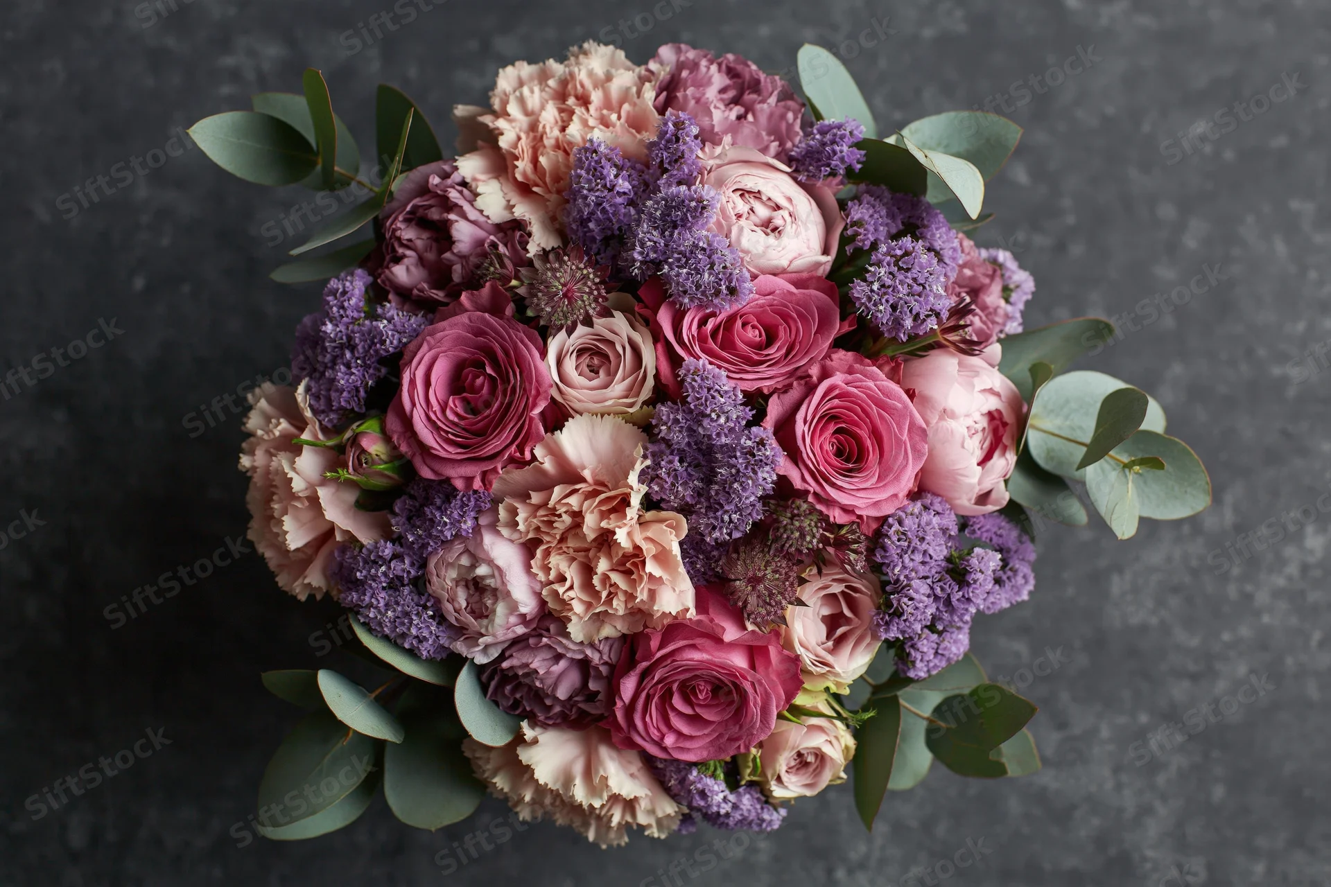Overhead close-up of a lush mixed flower bouquet with roses, peonies, carnations, lilac and eucalyptus on dark textured surface