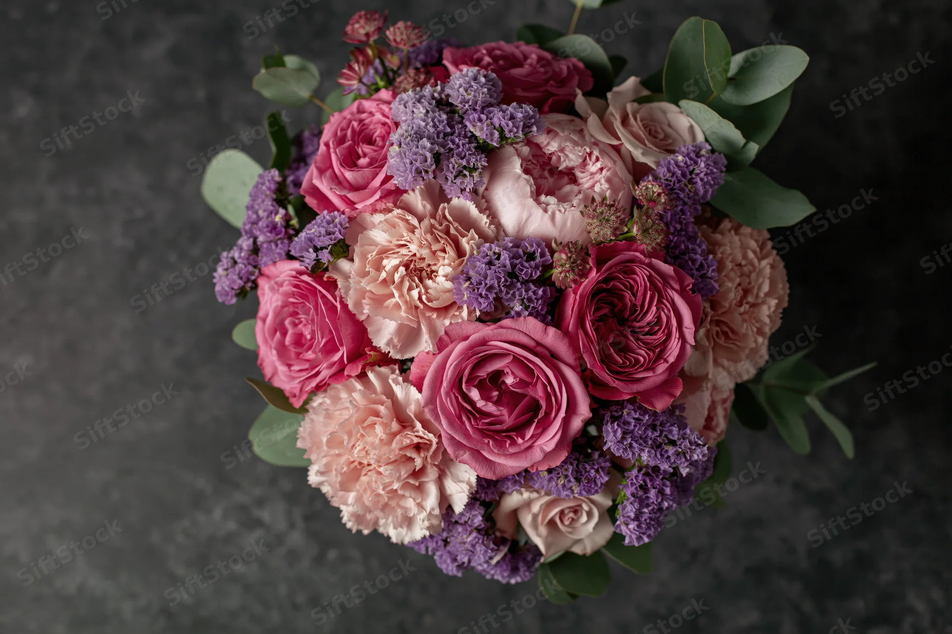 Overhead view of a lush mixed flower bouquet with pink roses, peonies, purple lilac, and green eucalyptus on dark textured surface