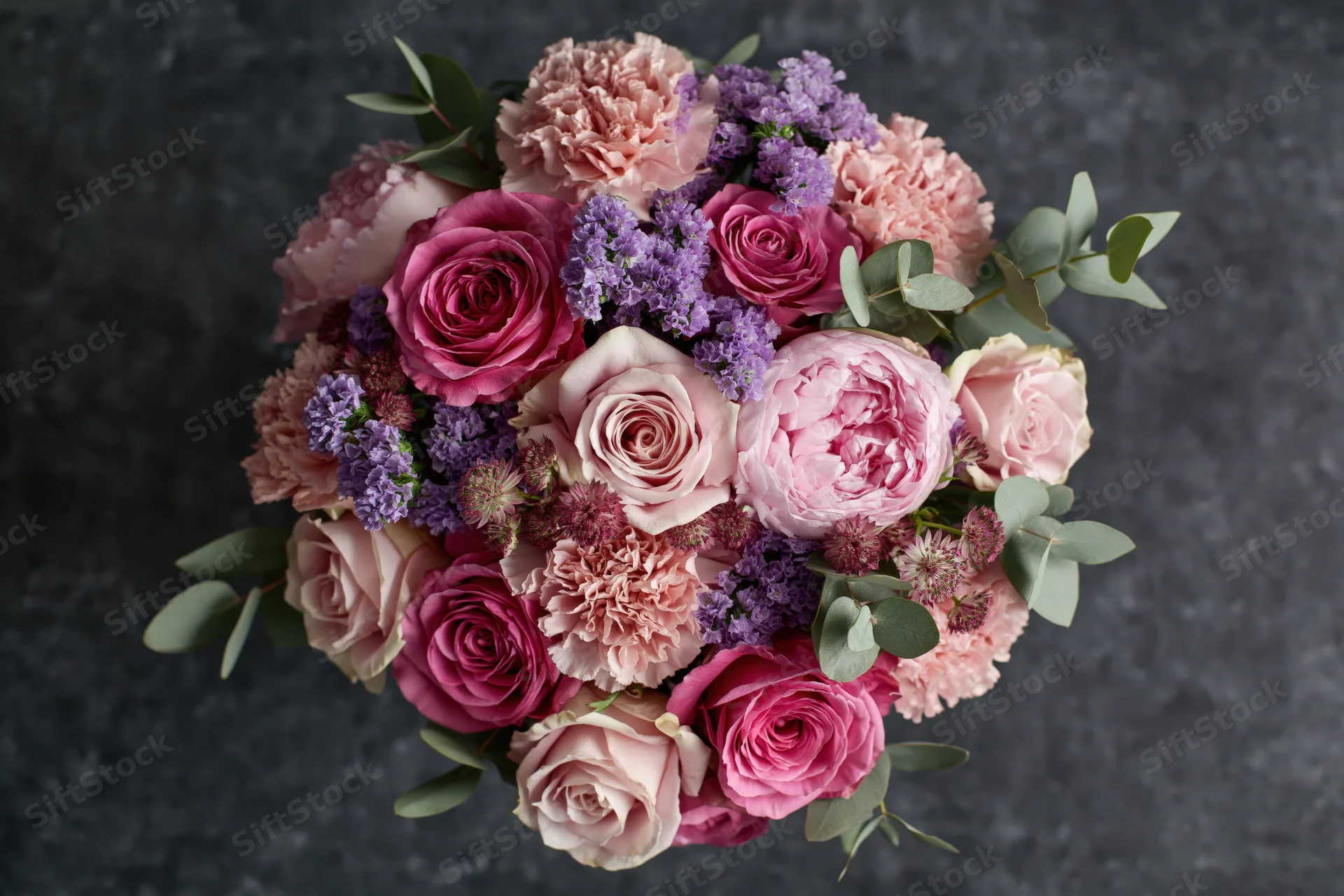 Overhead close-up of a lush mixed flower bouquet with pink roses, peonies, carnations, lavender and eucalyptus on dark textured surface