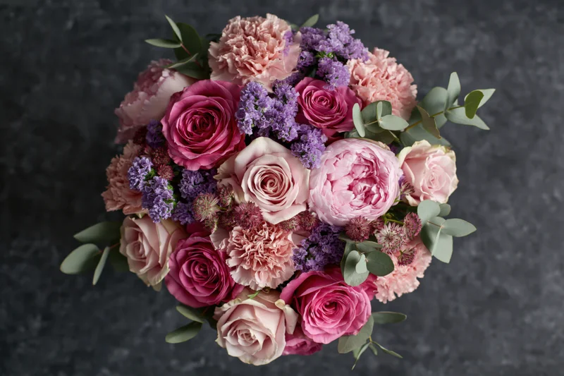 Overhead close-up of a lush mixed flower bouquet with pink roses, peonies, carnations, lavender and eucalyptus on dark textured surface