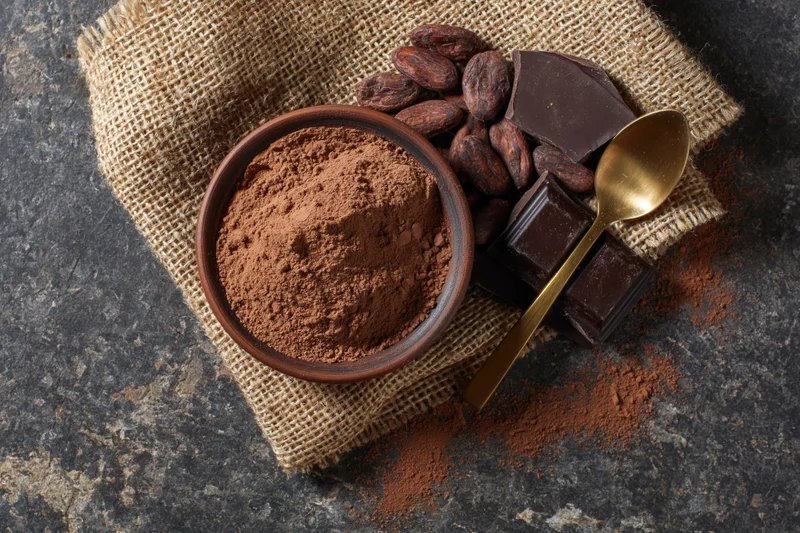 Cocoa powder in a clay bowl with cacao beans and chocolate pieces on burlap over stone surface
