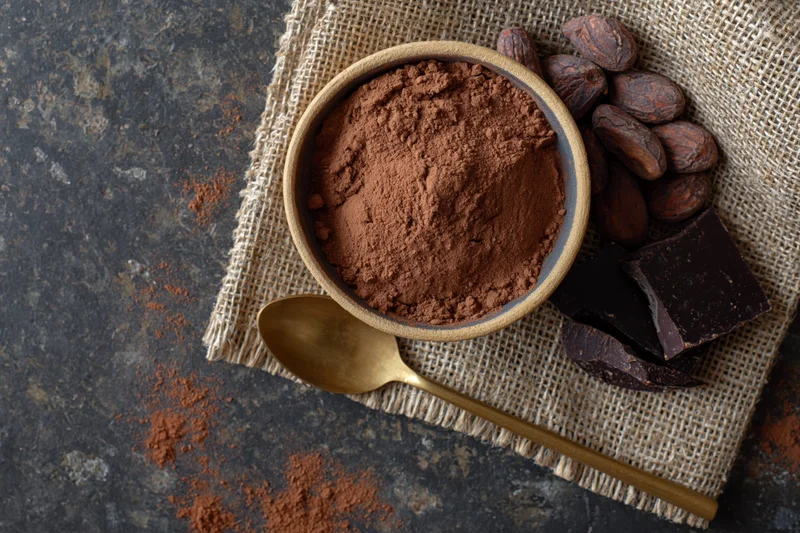 Cocoa powder in a clay bowl with cacao beans and chocolate pieces on burlap fabric