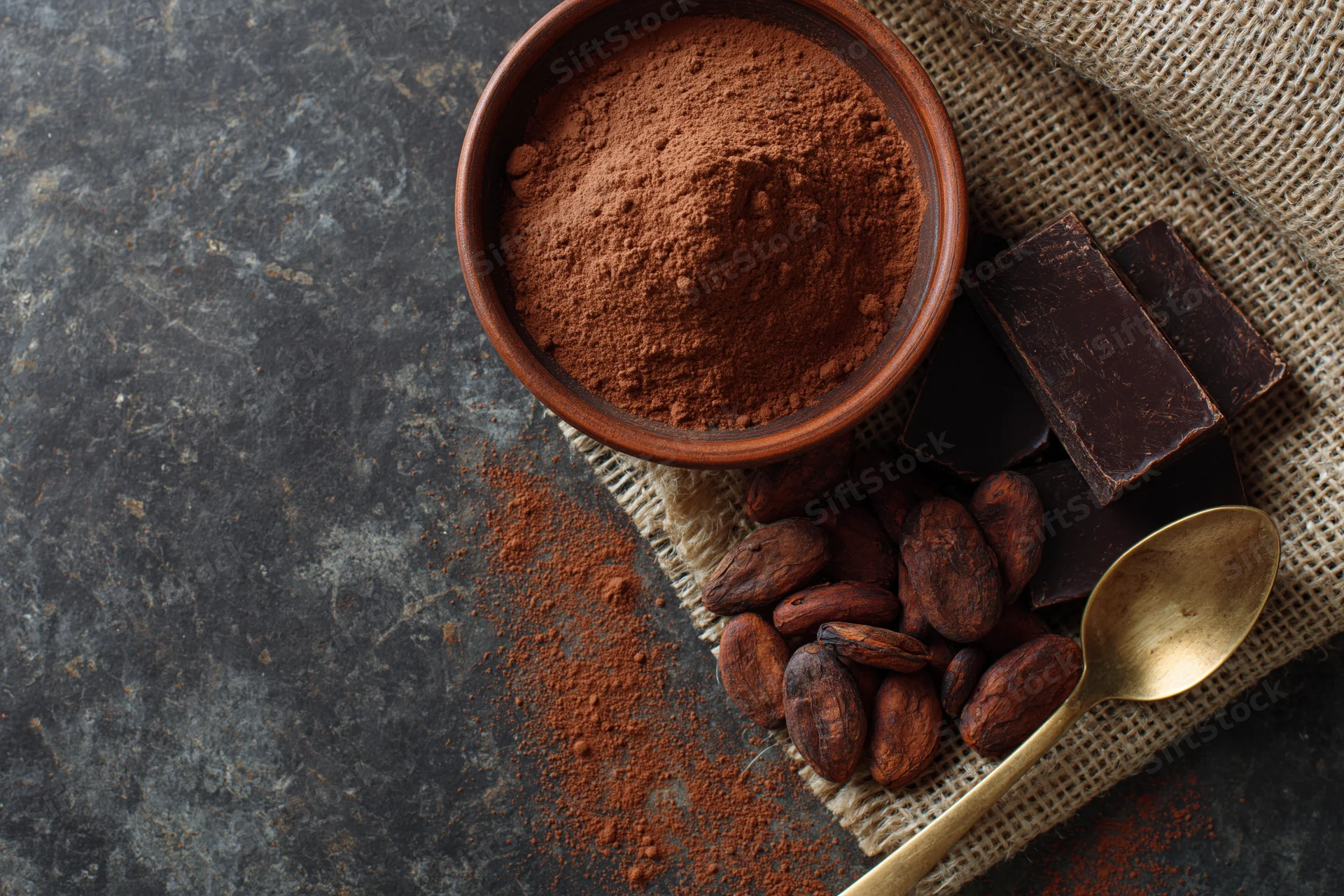 Cocoa powder in a clay bowl with chocolate bars, cocoa beans, and a spoon on burlap and stone surface