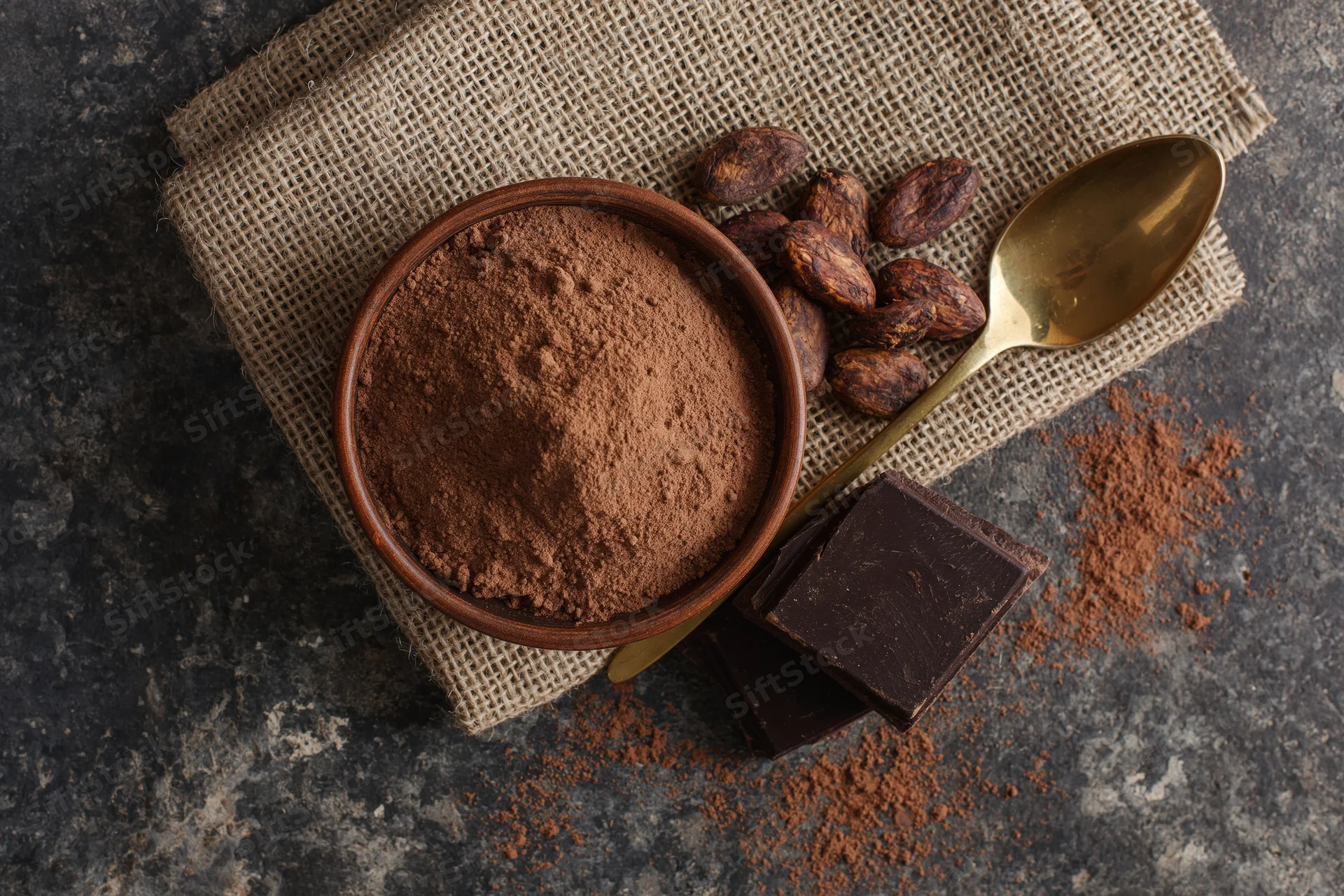 Cocoa powder in a clay bowl with roasted cacao beans and dark chocolate on burlap and stone surface