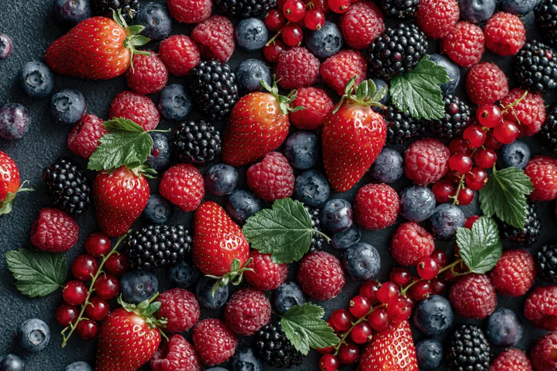 Overhead flat lay of mixed fresh berries densely arranged on dark surface with green leaves