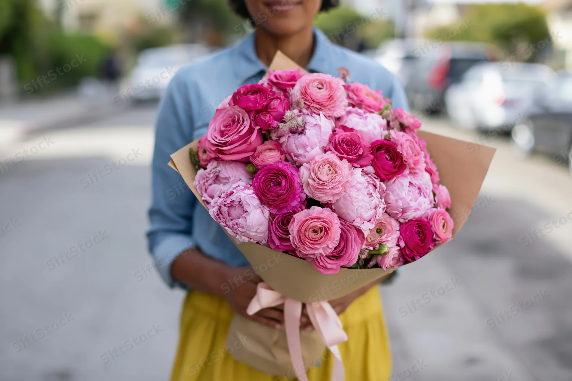 Woman holding a large bouquet of pink and magenta roses and peonies wrapped in brown paper on a street