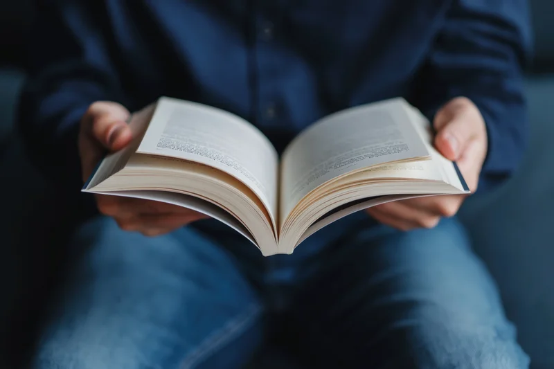 Person holding open paperback book with fanned pages while seated indoors