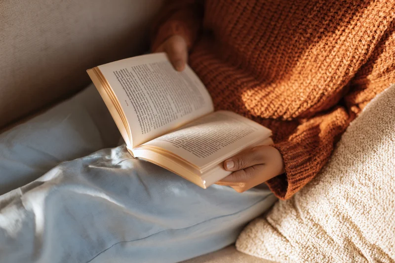 Woman reading an open paperback book on her lap while sitting on a couch