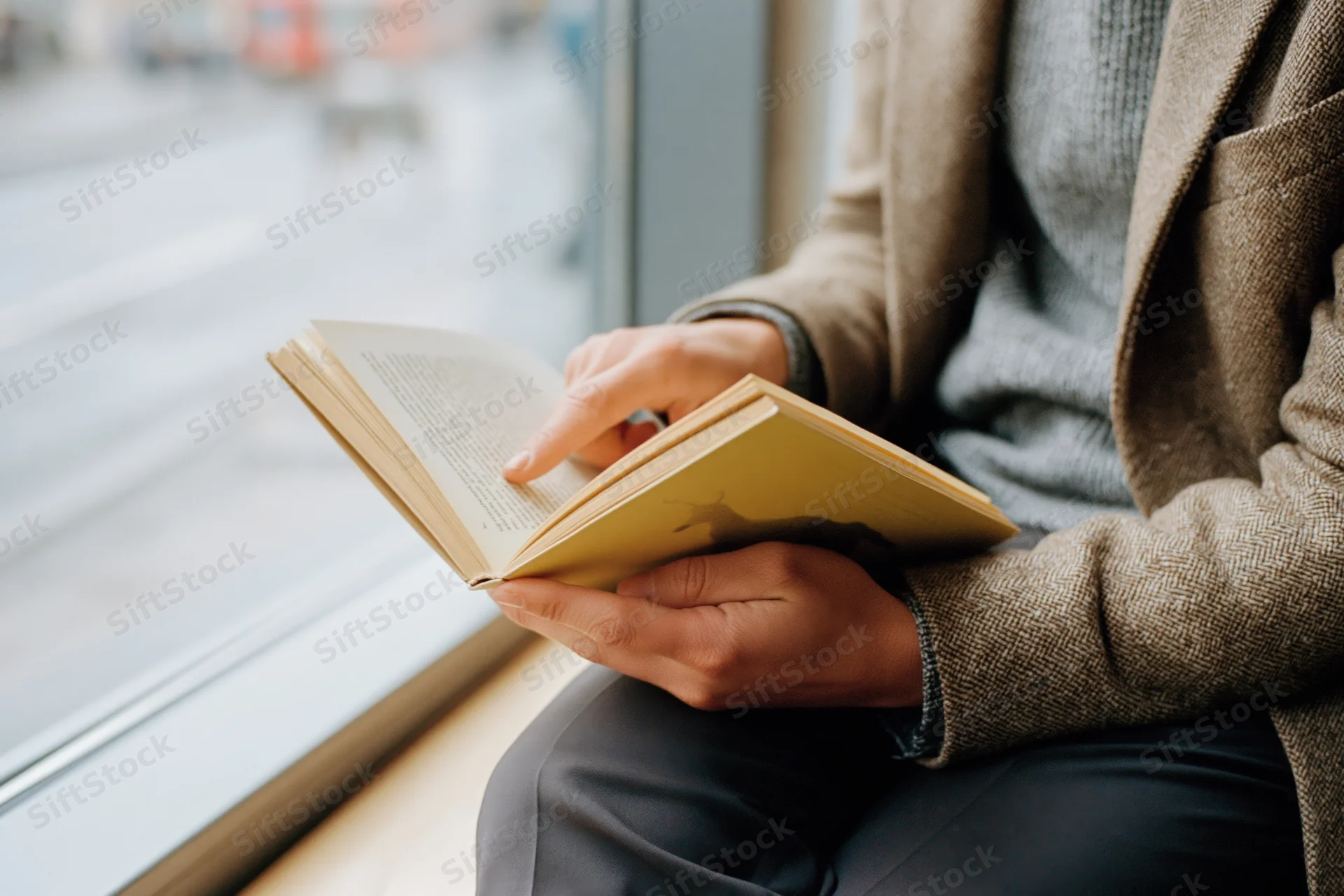 Woman reading a paperback book while sitting by a window indoors