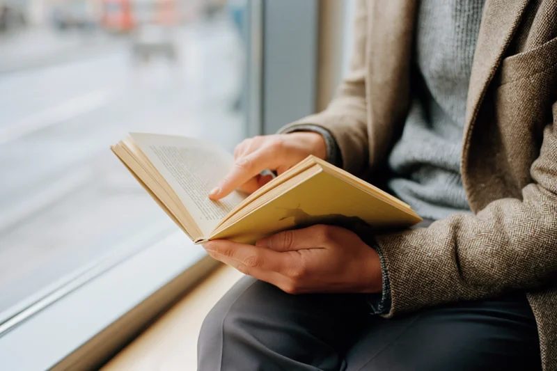 Woman reading a paperback book while sitting by a window indoors