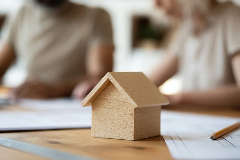 Wooden house model on a desk with blueprints and a couple in the background discussing plans