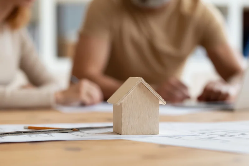 Wooden house model on blueprints with professionals in background discussing design