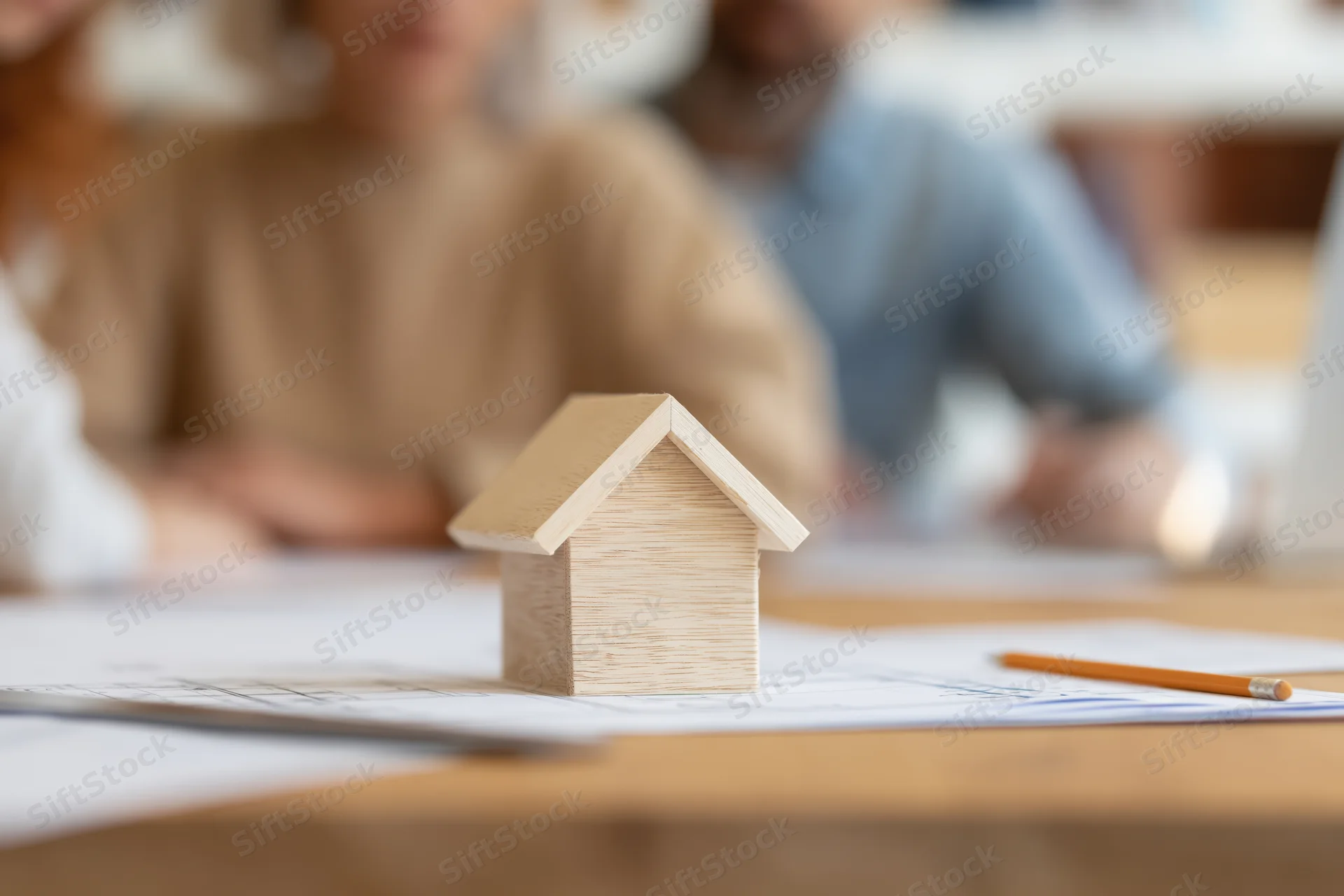 Wooden house model on blueprint plans on desk with professionals in background meeting