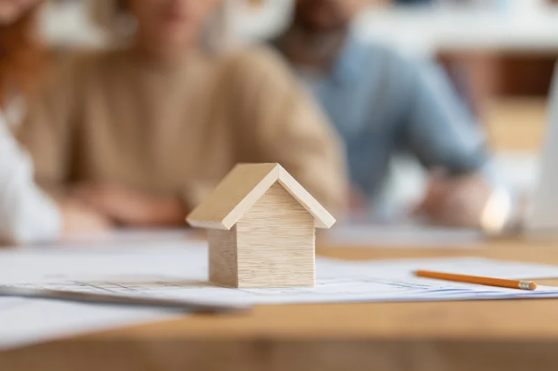 Wooden house model on blueprint plans on desk with professionals in background meeting