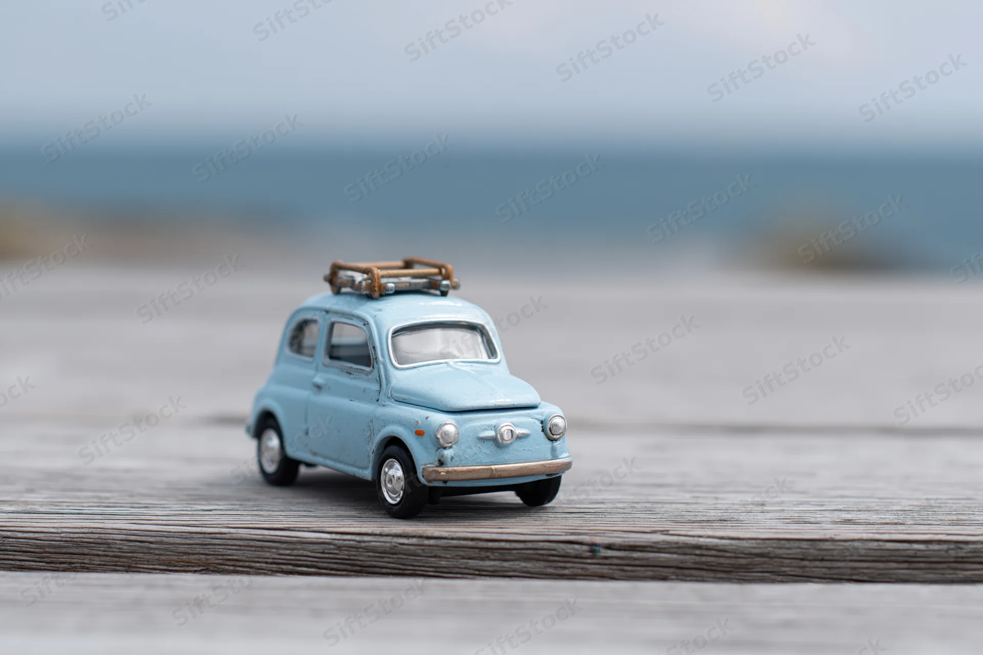 Small blue vintage toy car with a wooden luggage rack on a weathered wooden deck overlooking the ocean