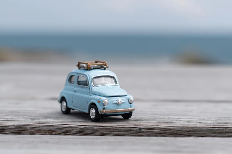 Small blue vintage toy car with a wooden luggage rack on a weathered wooden deck overlooking the ocean