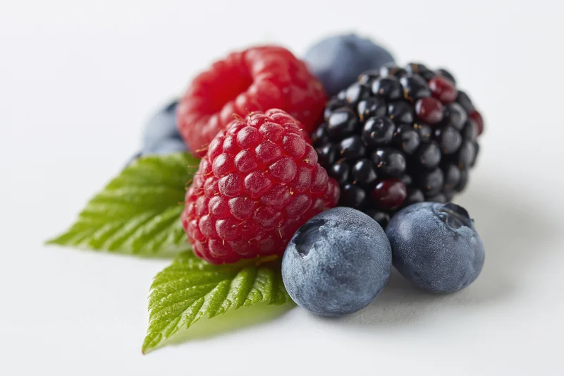 Cluster of fresh raspberries, blackberries and blueberries with green leaves on white surface