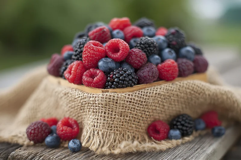 Wooden basket overflowing with fresh raspberries, blueberries and blackberries on burlap cloth over wooden surface